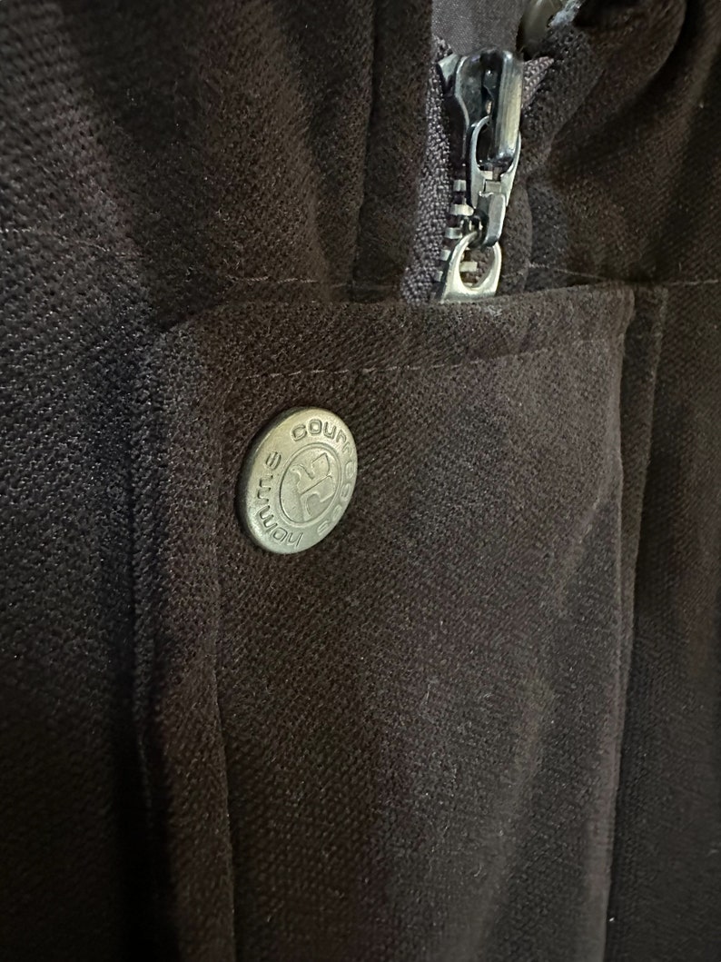 May include: A close-up of a brown zipper on a jacket with a metal button that says "Course" on it.