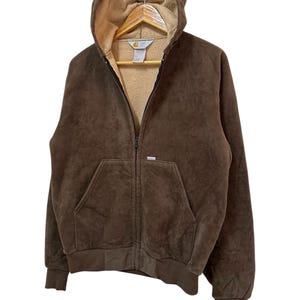 Puede incluir: Una sudadera con capucha de cremallera de color marr&oacute;n con capucha forrada de forro polar beige. La sudadera tiene un bolsillo canguro y un logotipo de Carhartt en el pecho izquierdo.