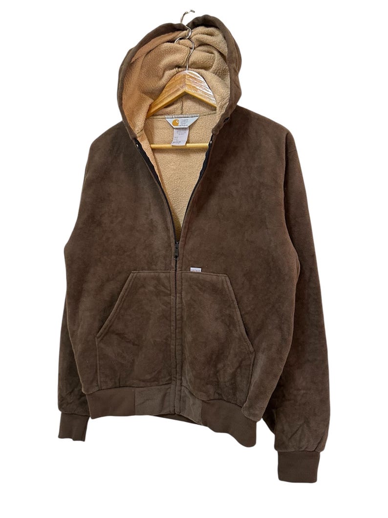 Puede incluir: Una sudadera con capucha de cremallera de color marr&oacute;n con capucha forrada de forro polar beige. La sudadera tiene un bolsillo canguro y un logotipo de Carhartt en el pecho izquierdo.