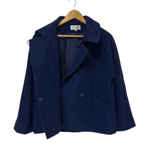 May include: A navy blue wool coat with a double-breasted design, featuring two buttons and two pockets. The coat is hanging on a wooden hanger against a white background. The label reads "HANAE MORI".