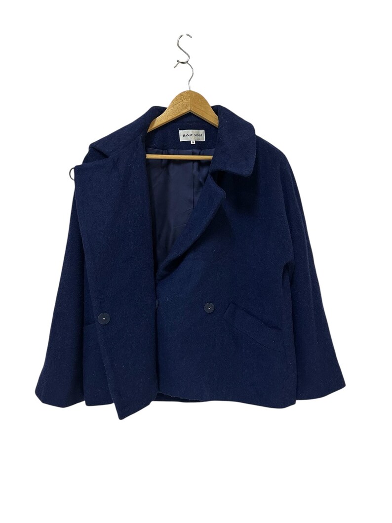 May include: A navy blue wool coat with a double-breasted design, featuring two buttons and two pockets. The coat is hanging on a wooden hanger against a white background. The label reads "HANAE MORI".