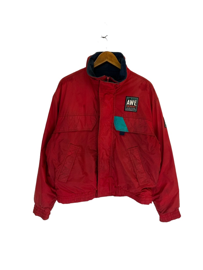 May include: Red zip-up jacket with a blue and green logo that says "AWE" on the front. The jacket has a high collar and a zipper closure.