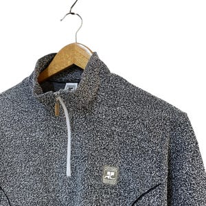 May include: A gray and white flecked zip-up sweater with a high collar. The sweater has a small logo patch on the chest and a white zipper. The garment is hanging on a wooden hanger.