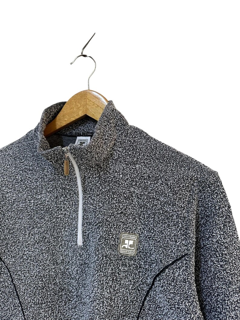May include: A gray and white flecked zip-up sweater with a high collar. The sweater has a small logo patch on the chest and a white zipper. The garment is hanging on a wooden hanger.