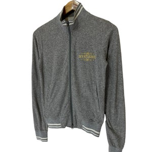 May include: A gray zip-up sweatshirt with white ribbed trim on the collar, cuffs, and hem. The sweatshirt has a gold embroidered logo that says "HYSTERIC" on the front.
