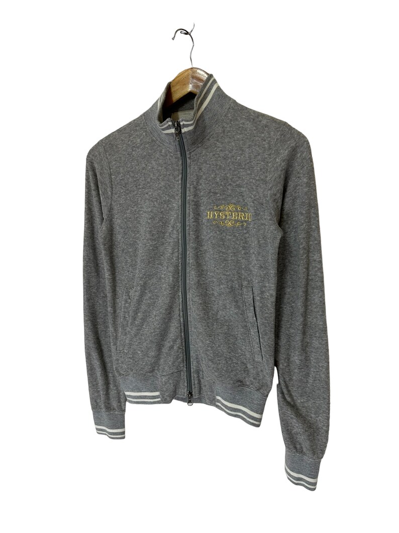 May include: A gray zip-up sweatshirt with white ribbed trim on the collar, cuffs, and hem. The sweatshirt has a gold embroidered logo that says "HYSTERIC" on the front.
