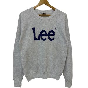 Può includere: Una felpa grigio mélange con la scritta "Lee" in blu navy sul petto. La felpa ha un girocollo e maniche lunghe. Il logo Lee è in un carattere in grassetto. Il capo è appeso a una gruccia di legno.