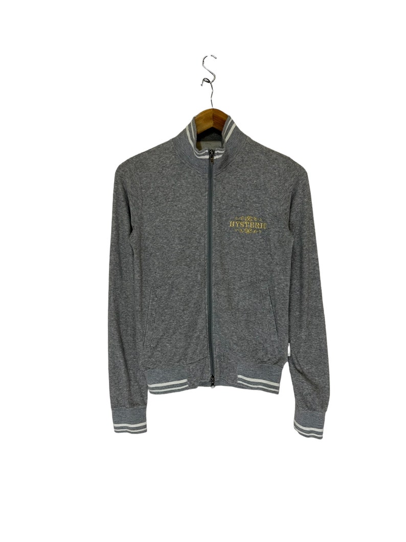 May include: A gray zip-up jacket with white ribbed trim on the cuffs and waistband. The jacket has a gold embroidered logo that says "HYSTERIC" on the front.