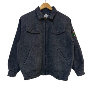 May include: A charcoal gray jacket with a full zipper closure, two chest pockets, and two lower pockets. The jacket has a small patch on the upper left sleeve and a ribbed hem. The jacket is hanging on a wooden hanger.