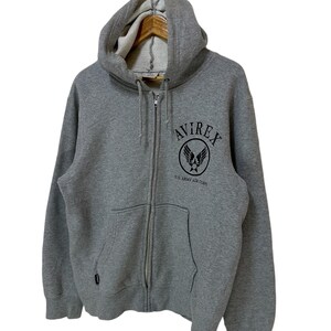 May include: A gray zip-up hoodie with a black embroidered logo that says "AVIREX" and "U.S. ARMY AIR CORPS".