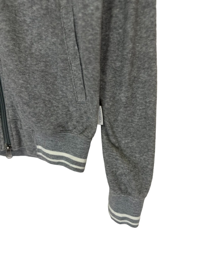 May include: A gray zip-up sweatshirt with white ribbed trim around the cuffs and bottom hem. The sweatshirt has a zipper closure and a pocket on the left side.