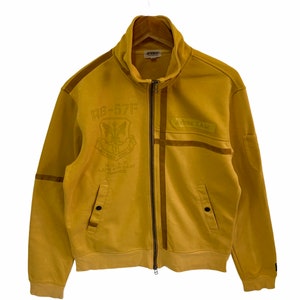Könnte beinhalten: Eine gelbe Reißverschlussjacke mit einem braunen Patch auf der rechten Seite, auf dem "Wrex T.A.W." steht. Die Jacke hat einen großen gestickten Patch auf der linken Seite, auf dem "RB-57F" und "U.S.A.F. LAON AIR BASE FRANCE" steht.
