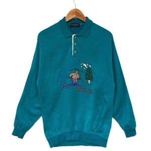 Puede incluir: Camiseta polo de manga larga azul turquesa con cuello blanco y cierre de botones. La camiseta presenta un diseño bordado de un esquiador en una escena de invierno con el texto "Ski Club" y "The Resort Club Vysoké Tatry".