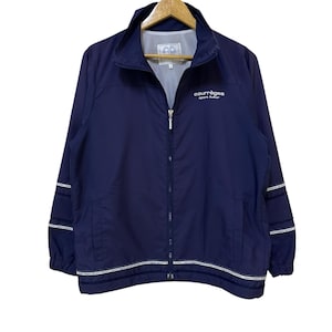 May include: Navy blue zip-up jacket with white trim and the text "courr&egrave;ges sport futur". The jacket has a stand-up collar, two front pockets, and white stripes on the sleeves. The jacket is hanging on a wooden hanger.
