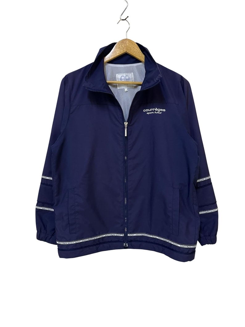 May include: Navy blue zip-up jacket with white trim and the text "courr&egrave;ges sport futur". The jacket has a stand-up collar, two front pockets, and white stripes on the sleeves. The jacket is hanging on a wooden hanger.