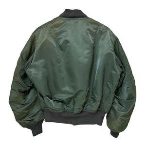 Puede incluir: Vista trasera de una chaqueta bomber verde con cuello, pu&ntilde;os y cintura de color gris oscuro. La chaqueta tiene una textura brillante y suave y una silueta cl&aacute;sica de bombardero. Los pu&ntilde;os y la cintura son acanalados.