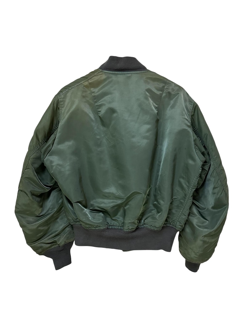 Puede incluir: Vista trasera de una chaqueta bomber verde con cuello, pu&ntilde;os y cintura de color gris oscuro. La chaqueta tiene una textura brillante y suave y una silueta cl&aacute;sica de bombardero. Los pu&ntilde;os y la cintura son acanalados.