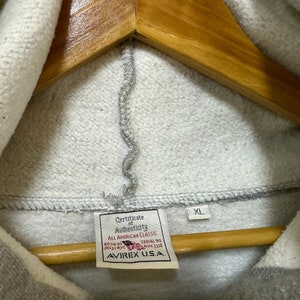 May include: A gray hooded sweatshirt with a zipper closure. The tag on the sweatshirt reads "Certificate of Authenticity, All American Classic, No:74-03 JRC32 NYC, Serial No. AVX 3328, AVIREX U.S.A." and "XL".