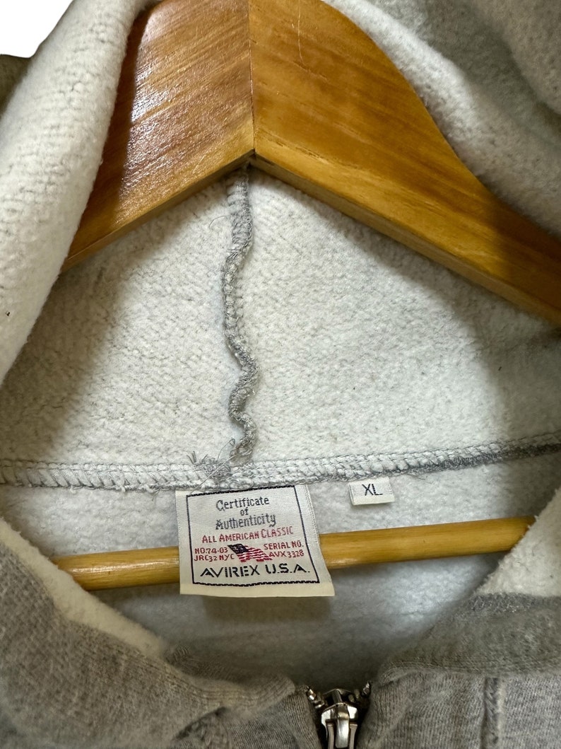 May include: A gray hooded sweatshirt with a zipper closure. The tag on the sweatshirt reads "Certificate of Authenticity, All American Classic, No:74-03 JRC32 NYC, Serial No. AVX 3328, AVIREX U.S.A." and "XL".