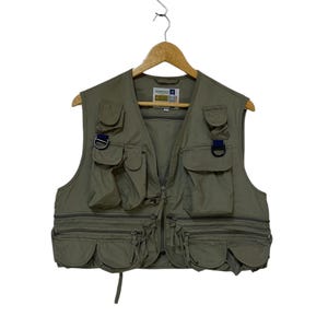 Puede incluir: Chaleco de pesca verde oliva con múltiples bolsillos y cremalleras. El chaleco tiene una cremallera frontal y un detalle de anilla en D. El chaleco está colgado de una percha de madera.