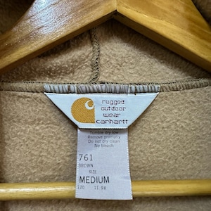 Puede incluir: Sudadera con capucha Carhartt marr&oacute;n con una etiqueta que dice "rugged outdoor wear Carhartt" y "Tumble dry low Remove promptly Do not dry clean No bleach" y "761 BROWN SIZE MEDIUM 120 11 98".