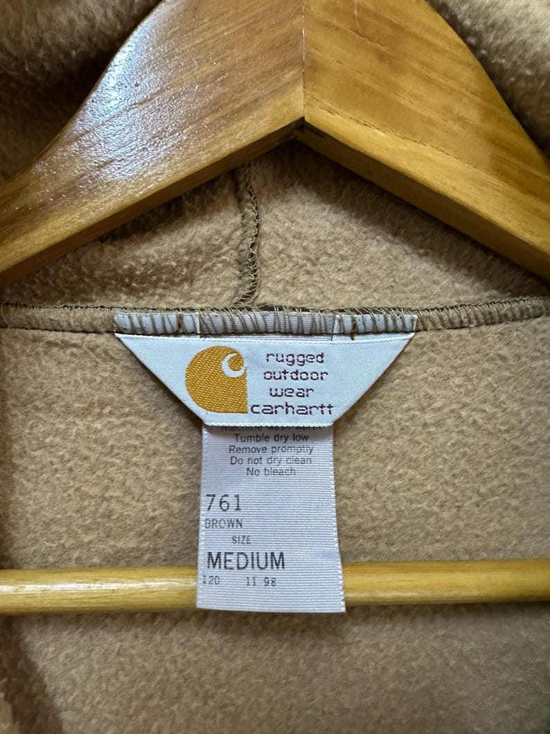 Puede incluir: Sudadera con capucha Carhartt marr&oacute;n con una etiqueta que dice "rugged outdoor wear Carhartt" y "Tumble dry low Remove promptly Do not dry clean No bleach" y "761 BROWN SIZE MEDIUM 120 11 98".