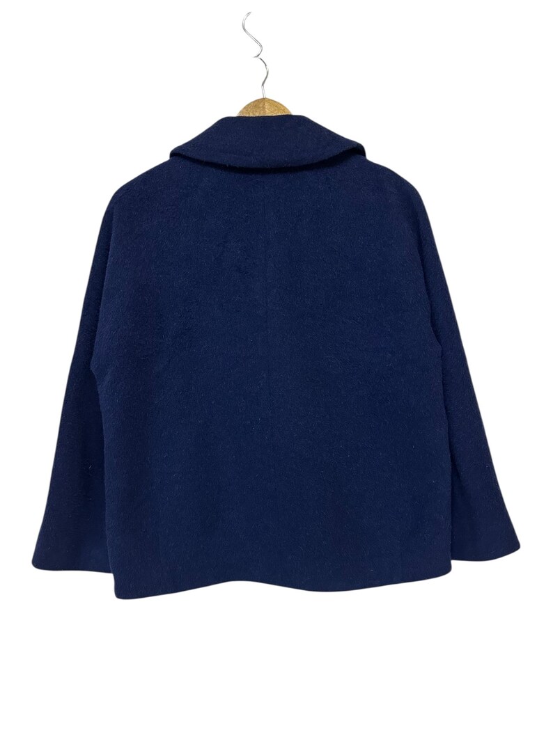 May include: A navy blue wool coat with a wide collar, hanging on a wooden hanger. The back of the coat is visible, showcasing its simple, classic design. The coat's fabric appears soft and has a slight texture.