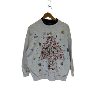 Puede incluir: Un suéter navideño gris con un cuello interior negro. El suéter presenta un gran diseño de árbol de Navidad hecho de pequeñas figuras de Papá Noel. El árbol está rodeado de copos de nieve, estrellas y notas musicales. El texto "Can find Rudolph?" está impreso en la parte inferior del árbol.