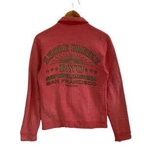 May include: A red long-sleeve shirt with a gold embroidered logo that says "Shore Breeze Ryu Surf Rider, California San Francisco Since 1979".