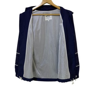 May include: Navy blue hooded jacket with a white interior lining, hanging on a wooden hanger. The jacket has a zipper closure and white drawstrings at the bottom. The jacket has white accents on the sleeves and a small logo on the inside.