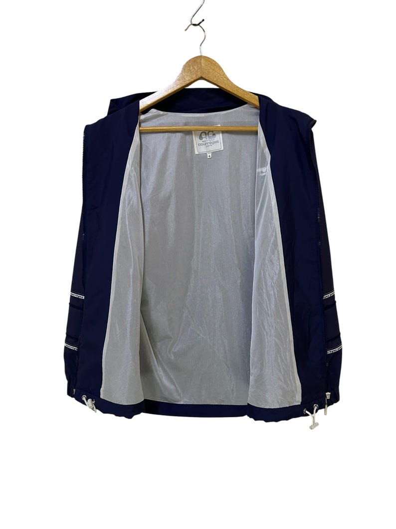 May include: Navy blue hooded jacket with a white interior lining, hanging on a wooden hanger. The jacket has a zipper closure and white drawstrings at the bottom. The jacket has white accents on the sleeves and a small logo on the inside.