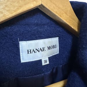 May include: Close-up of a navy blue coat with the label "HANAE MORI" visible. The label also shows the size "38". The coat is hanging on a wooden hanger.