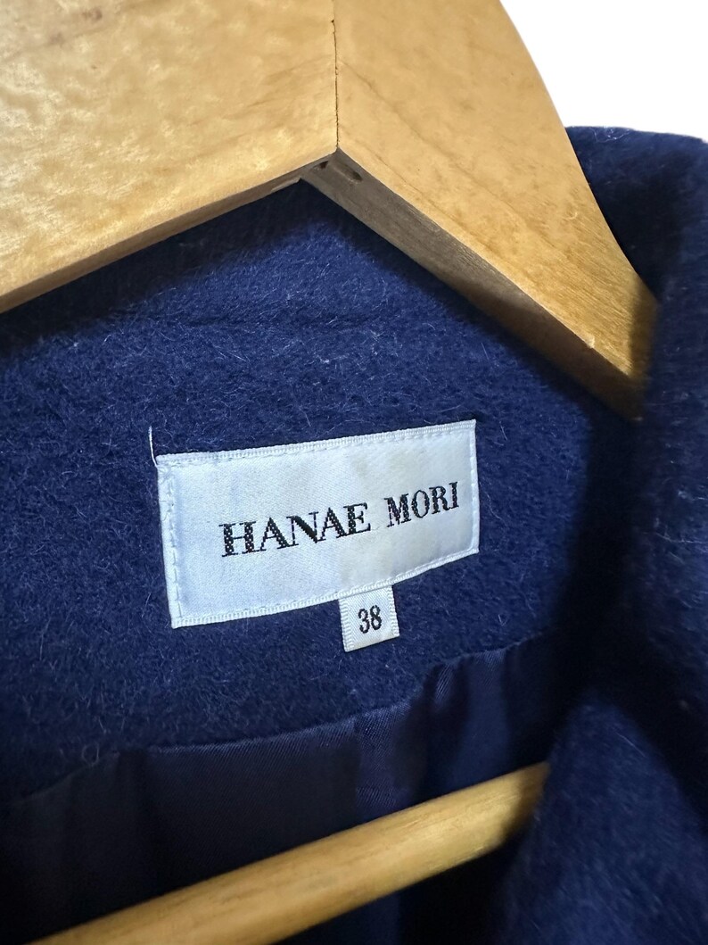 May include: Close-up of a navy blue coat with the label "HANAE MORI" visible. The label also shows the size "38". The coat is hanging on a wooden hanger.