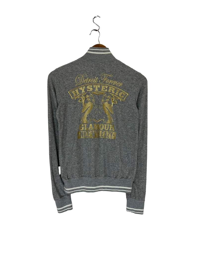 May include: Gray long-sleeve sweatshirt with a white ribbed collar and cuffs. The back of the sweatshirt features a gold embroidered design that says "Detroit Forever Hysteric Glamour 1972".