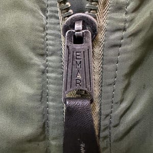 Puede incluir: Primer plano de una cremallera de chaqueta verde oliva con un tirador met&aacute;lico. El tirador est&aacute; grabado con la palabra "EMAR" y tiene un tirador de cuero negro. Los dientes de la cremallera son de color met&aacute;lico a juego.