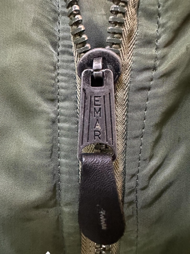 Puede incluir: Primer plano de una cremallera de chaqueta verde oliva con un tirador met&aacute;lico. El tirador est&aacute; grabado con la palabra "EMAR" y tiene un tirador de cuero negro. Los dientes de la cremallera son de color met&aacute;lico a juego.