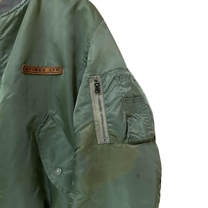 Puede incluir: Chaqueta bomber verde oliva con cuello y pu&ntilde;os grises. Cuenta con un bolsillo con cremallera en la manga izquierda y un peque&ntilde;o parche con el texto "AVIREX LTD". La chaqueta tiene un estilo bomber cl&aacute;sico.