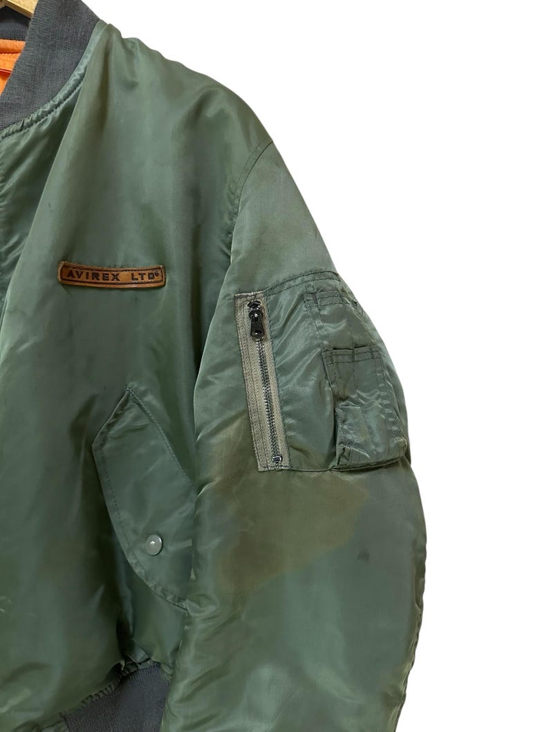 Puede incluir: Chaqueta bomber verde oliva con cuello y pu&ntilde;os grises. Cuenta con un bolsillo con cremallera en la manga izquierda y un peque&ntilde;o parche con el texto "AVIREX LTD". La chaqueta tiene un estilo bomber cl&aacute;sico.