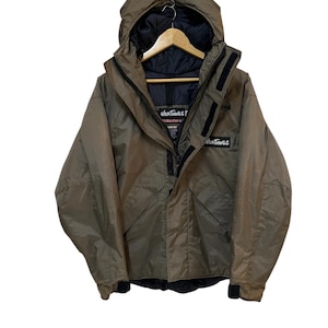May include: A tan Wild Things jacket with a hood, black lining, and the brand name on the chest. The jacket has a zipper closure and multiple pockets. The jacket is hanging on a wooden hanger.