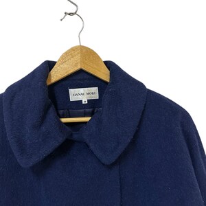 May include: A navy blue coat with a wide collar, hanging on a wooden hanger. The coat has the brand name "HANAE MORI" on a tag. The coat is made of a soft, textured fabric.