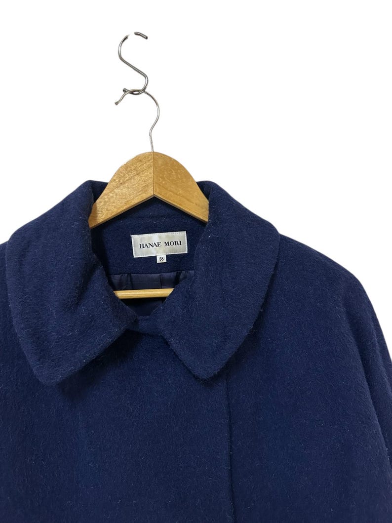 May include: A navy blue coat with a wide collar, hanging on a wooden hanger. The coat has the brand name "HANAE MORI" on a tag. The coat is made of a soft, textured fabric.