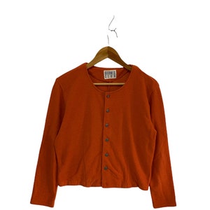 May include: A long-sleeved, orange button-up cardigan with a round neckline. The cardigan has a label that reads "Gitanes Club".