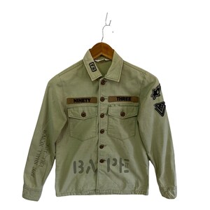 May include: A light green utility jacket with brown patches that say "Ninety" and "Three". The jacket has a black and white graphic on the front that says "BAPE" and the text "APE SHALL NEVER KILL APE" on the left sleeve.