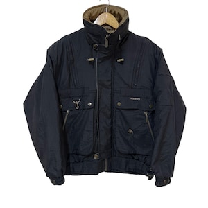 Puede incluir: Chaqueta negra vintage con cuello alto y múltiples bolsillos. La chaqueta tiene un forro marrón en el cuello y cierre de cremallera. La marca "DESCENTE" es visible en el lado derecho del pecho. La chaqueta está colgada en una percha.