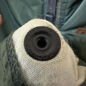 Puede incluir: Primer plano de una chaqueta verde oscuro con cierre de bot&oacute;n a presi&oacute;n negro. El bot&oacute;n a presi&oacute;n es circular con el texto "MFG. CO." en el borde. La chaqueta tiene una cremallera y un forro de tela clara.