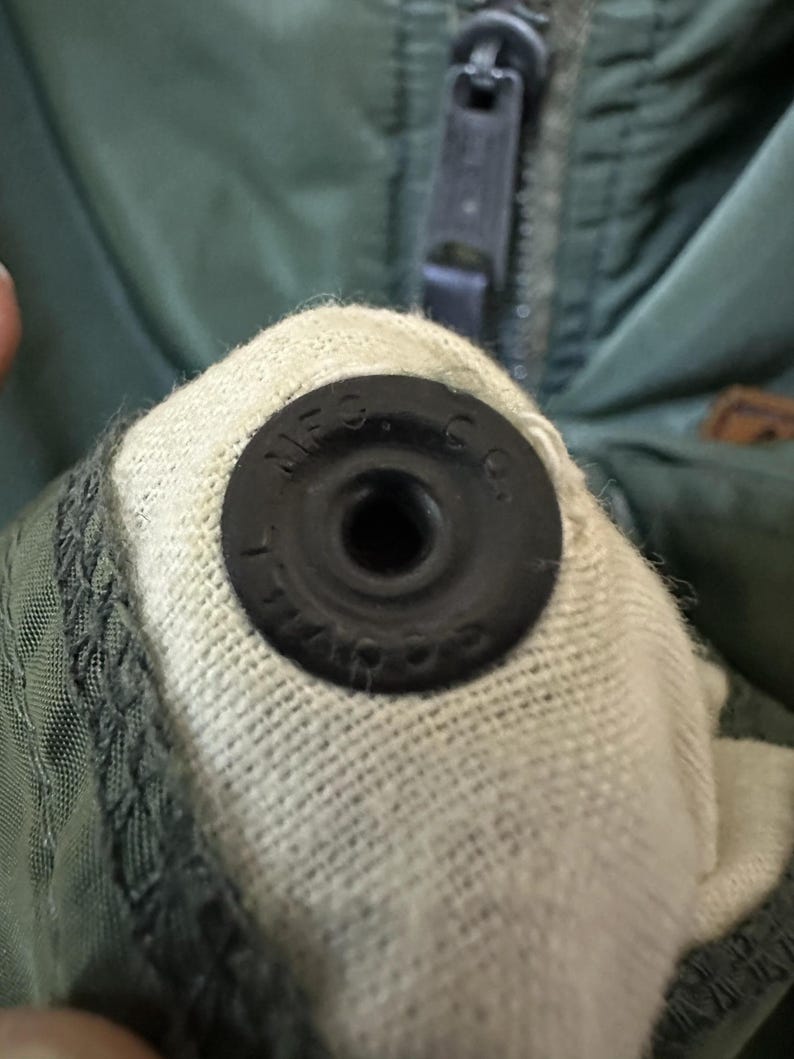Puede incluir: Primer plano de una chaqueta verde oscuro con cierre de bot&oacute;n a presi&oacute;n negro. El bot&oacute;n a presi&oacute;n es circular con el texto "MFG. CO." en el borde. La chaqueta tiene una cremallera y un forro de tela clara.