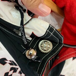 May include: Black and white jacket with a red lining. The jacket has a zipper closure and a silver button with the text "Sports" and a star. The jacket has a white drawstring with the text "Courages" printed on it.