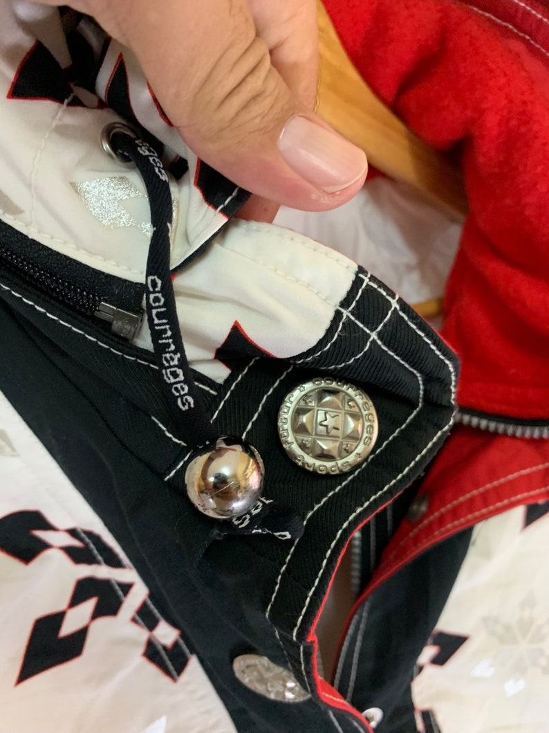 May include: Black and white jacket with a red lining. The jacket has a zipper closure and a silver button with the text "Sports" and a star. The jacket has a white drawstring with the text "Courages" printed on it.