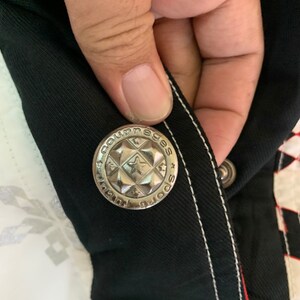 May include: Close-up of a silver button with the words "Sportswear" and "Courage" on it. The button is attached to a black fabric with white stitching.