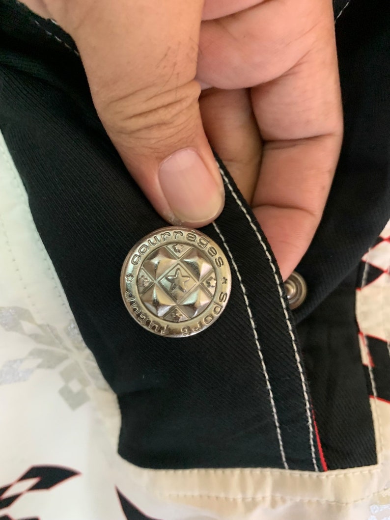 May include: Close-up of a silver button with the words "Sportswear" and "Courage" on it. The button is attached to a black fabric with white stitching.
